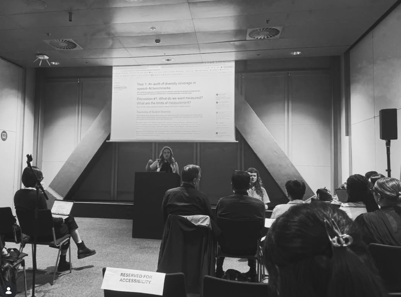 Maria Teleki presenting at a podium to a seated audience, with a slide on speech AI benchmarks behind her